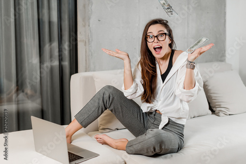 Fotografia Happy woman earning and saving money with laptop on couch at modern home