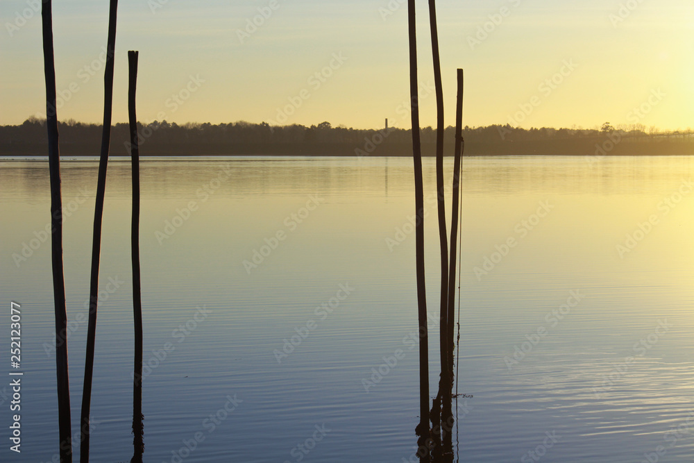 Reflect of sunset at the calm river, with wooden sticks on the first plan Reflect of sunset at the calm river, with wooden sticks on the first plan