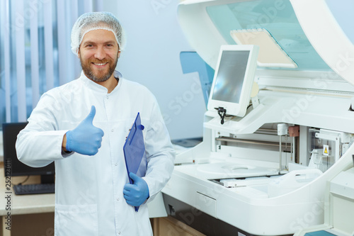 Handsome medical worker in lab coat standing near new equipment and showing like. Young doctor in blue gloves smiling at camera and holding finger up. Man working in laboratory in private clinic.