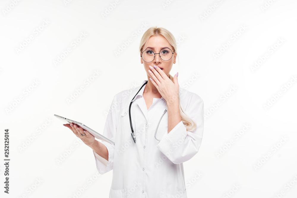 Portrait of young female blond doctor in white working uniform looking at camera isolated on white. A stethoscope is hanging on the neck. Closes the mouth with a hand in surprise. Portrait of young female blond doctor in white working uniform looking at camera isolated on white. A stethoscope is hanging on the neck. Closes the mouth with a hand in surprise.