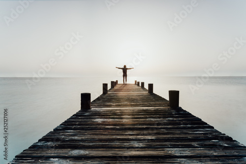 Person steht mit offenen Armen bei Sonnenuntergang auf einem Steg am Meer und genießt die Freiheit photo