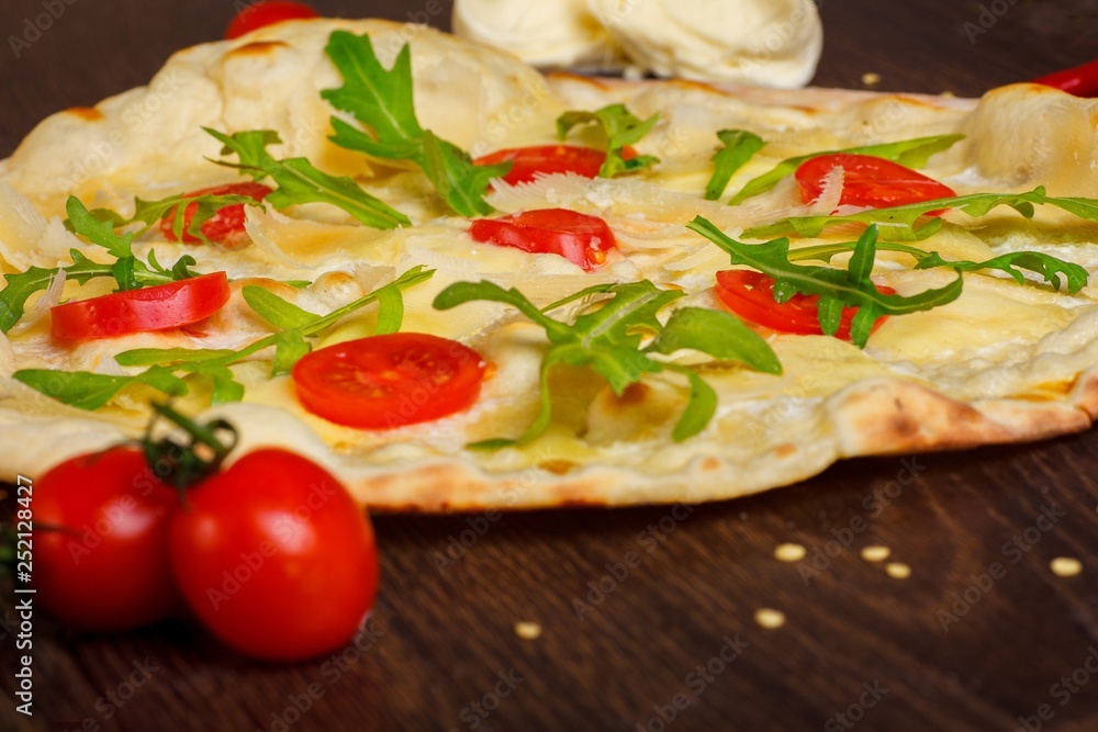 Vegetarian Italian pizza with melted mozzarella, parmezan cheese, red tomatoes and fresh green arugula leaves on a brown table decorated by mozzarella, red sweet pepper, cherry tomatoes and battledore
