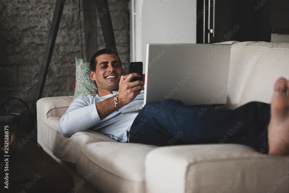 Happy young man lying on sofa at home using cell phone and laptop