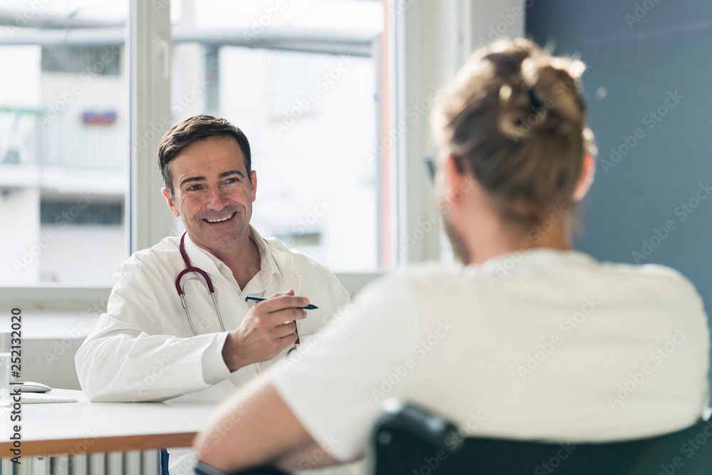 Smiling doctor talking to patient in wheelchair in medical practice Smiling doctor talking to patient in wheelchair in medical practice