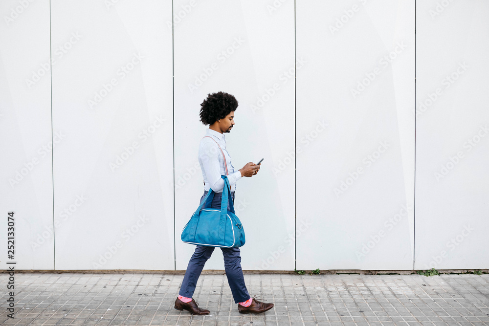 Man with bag walking in front of a white wall while looking at cell phone Man with bag walking in front of a white wall while looking at cell phone