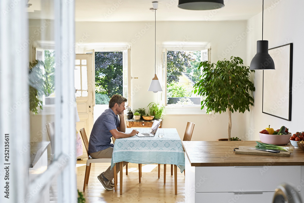 Mature man sitting in his dining room, using laptop Mature man sitting in his dining room, using laptop