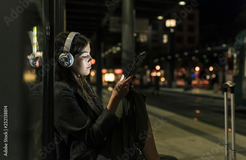 Young woman with headphones waiting at the station by night using tablet photo