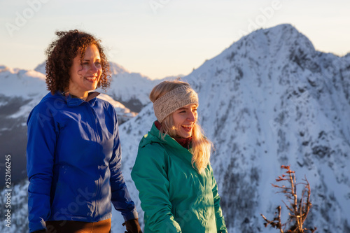 Couple friends are having fun on top of a mountain during a vibrant winter sunset. Taken on top of Zoa Peak near Hope, British Columbia, Canada.