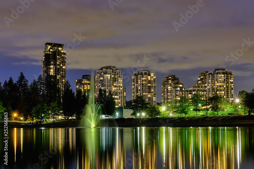 Night scape city view, Coquitlam, Greater Vancouver area, Canada. Fountain in lake and water reflections of modern buildings windows lights