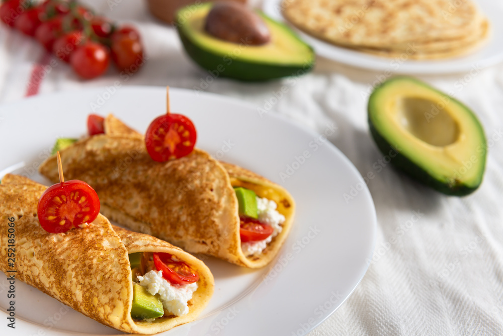 Crepe pancakes with avocado, soft white cheese and cherry tomatoes on white plate. Cherry tomatoes, avocado and stack of crepes on side. Close up