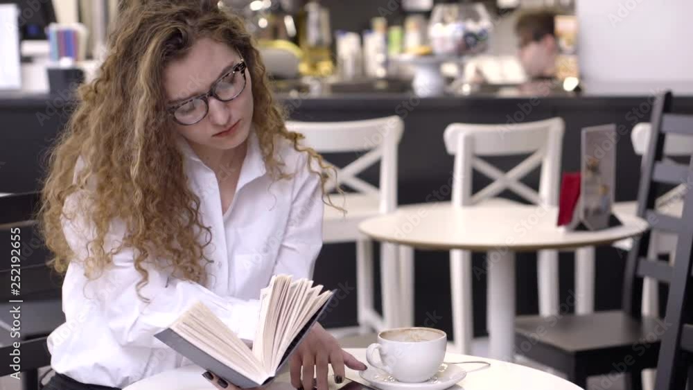 pretty woman is reading book, sitting at table in cafe, taking mobile phone and answering on sms pretty woman is reading book, sitting at table in cafe, taking mobile phone and answering on sms