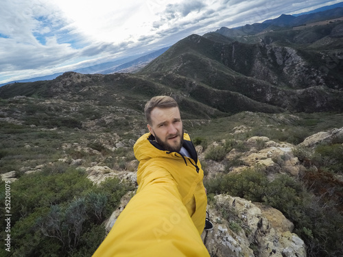 Young bearded male hiker takes a wide angle selfie with mountains in background.