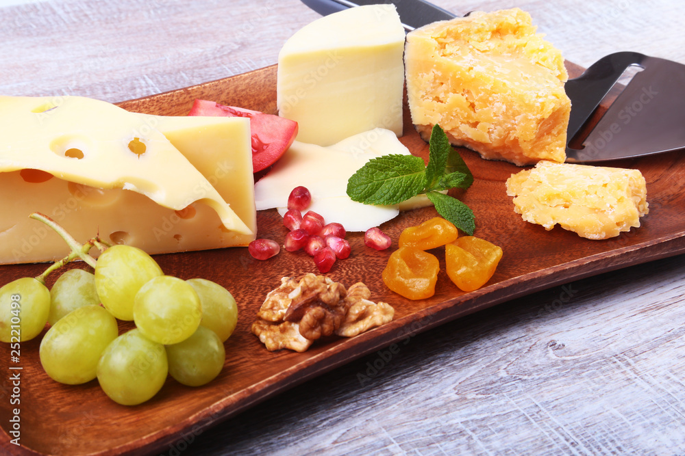 Assortment of cheese with fruits, grapes, nuts and cheese knife on a wooden serving tray.