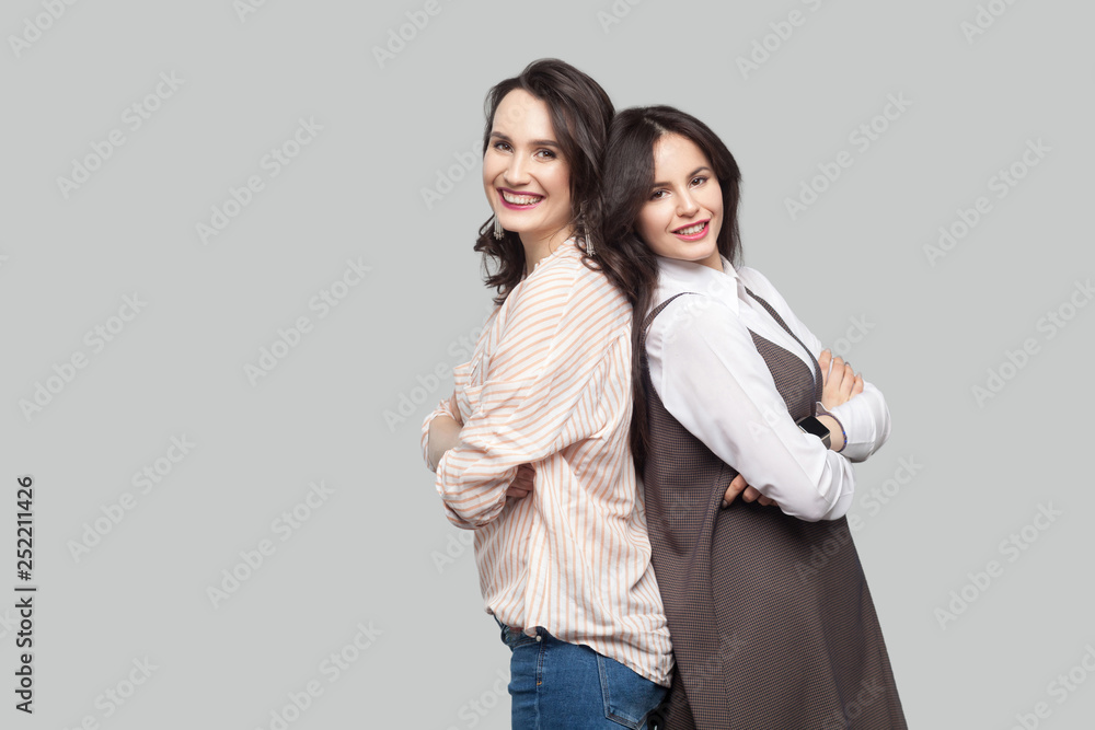 Portrait of two happy satisfied beautiful partners in casual style standing back to back with crossed arms and looking at camera with toothy smile. indoor studio shot, isolated on grey background. Portrait of two happy satisfied beautiful partners in casual style standing back to back with crossed arms and looking at camera with toothy smile. indoor studio shot, isolated on grey background.