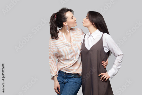 Portrait of two funny cheerful beautiful brunette best friends in casual style standing, hugging and looking at each other and kissing. indoor studio shot, isolated on grey background.