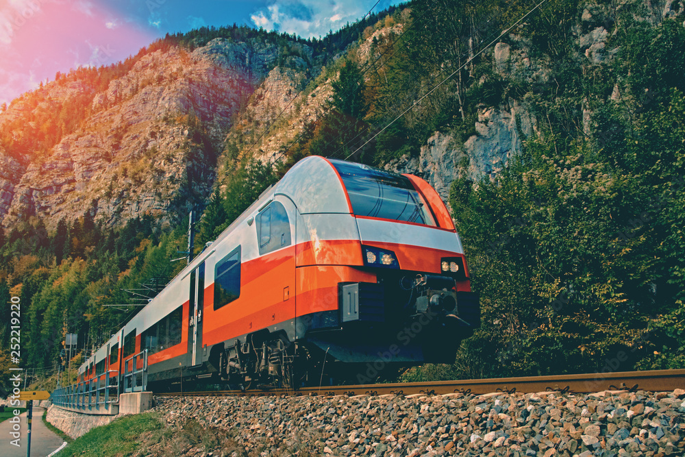 Obraz premium Red blue train in motion in Austrian alps mountains. High speed mountain train arrives at Hallstatt Obertraun train station in mountains. Location: resort village Hallstatt, Salzkammergut, Austria