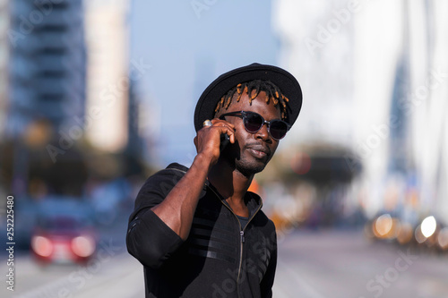 Front view of black man with sunglasses and hat standing against cityscape on the street while using a mobile phone in sunny day.