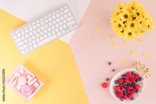 Flat lay and view from above of work office desk with labtop keyboard, flowers and berries on pink and yellow background. Layout of summer female pastel table