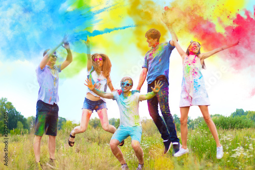 Cheerful and happy friends soiled by bright paints jumping and laugh in colorful smoke on nature. Company of young people having fun with holi paints on spring summer festival. Holi party concept.