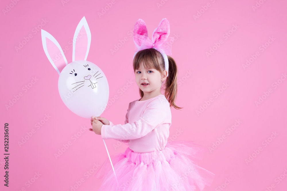 Cute pink young girl child daughter wears pink dress like rabbit playing with rabbit balloon with rabbit ears on isolated pink background. Spring is coming and Easter holiday concept