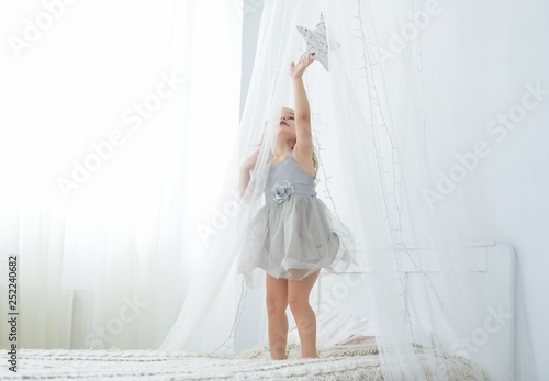 Little toddler girl standing on bed in white bedroom. Cute baby in nice dress tuching a star. photo