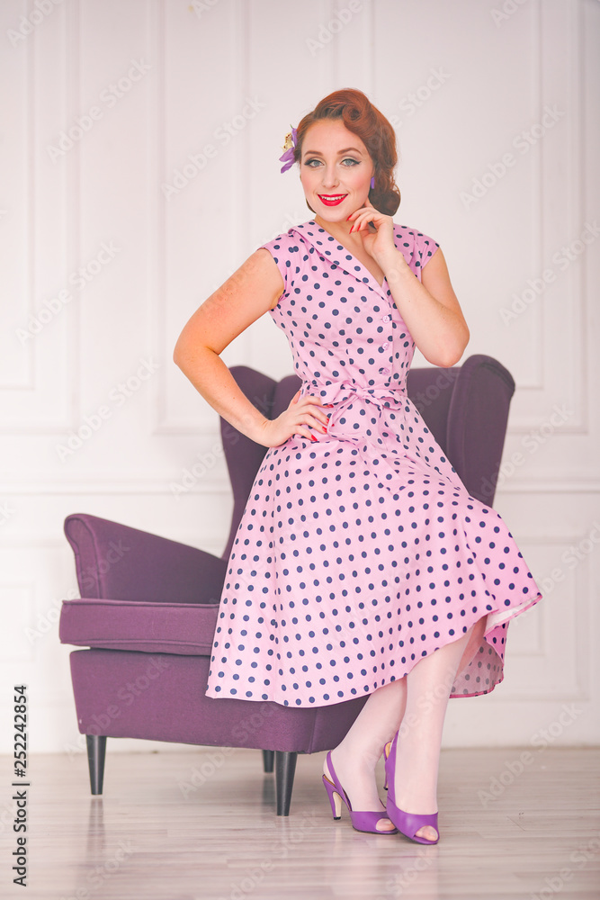 pretty redheaded pin up woman wearing pink polka dot dress and posing with purple armchair on white background pretty redheaded pin up woman wearing pink polka dot dress and posing with purple armchair on white background