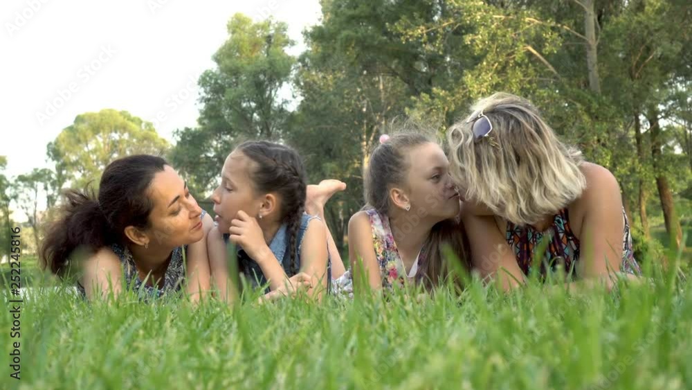 Two mothers and two little daughters lie on the green grass in the Park at sunset, playing and kissing. Portrait. Close up. View from ground level. 4K. 25 fps. Two mothers and two little daughters lie on the green grass in the Park at sunset, playing and kissing. Portrait. Close up. View from ground level. 4K. 25 fps.
