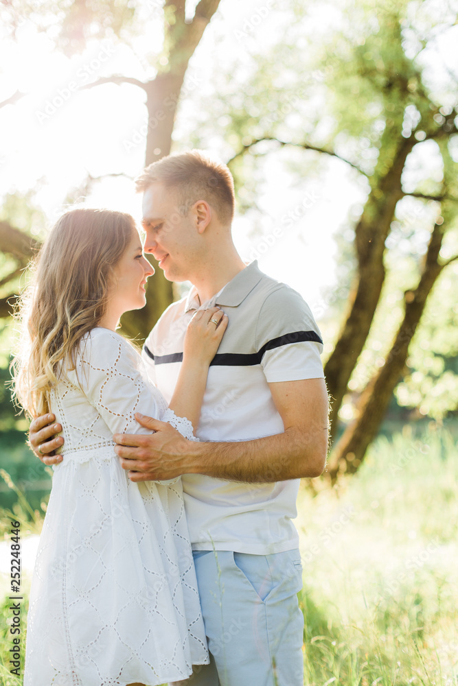 Fototapeta premium Young pretty couple in love standing in park. Handsome cheerful blonde girl in white dress hugging her boyfriend. Man and woman having fun outdoors
