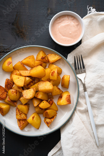 Plate of hot Patatas Bravas, a traditional Spanish tapas dish, fried or oven baked potato cubes until crispy served with spicy aioli garlic mayonnaise with chili and paprika on dark wooden background