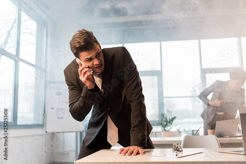 frightened businessman talking on smartphone in office with smoke near female coworker