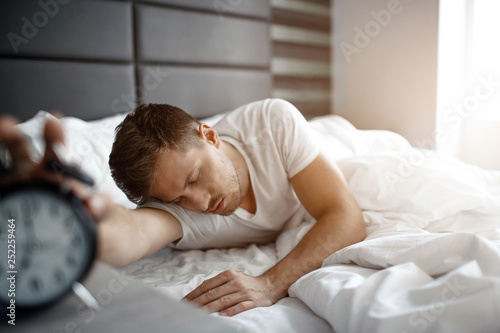 Young man sleep on bed in morning. He hold hand on clock. Oversleep. Daylight.