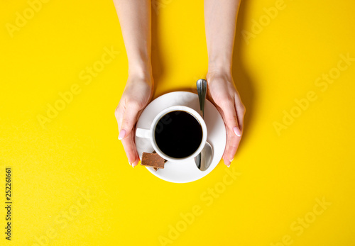 Cup of coffee with chocolate pieces in hand on yellow background, top view