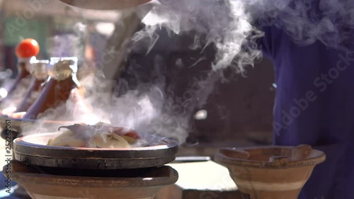Traditional moroccan Tajine food cooking in Tajine pots on the fire with smoke and tomatoes on top. Hand of local Cook is opening boiling dish and release smoke at bright sunny weather