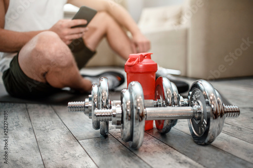Cut view of young well-built man go in for sports in apartment. He sit on floor and hold phone. Dumbbells on floor in front of man.
