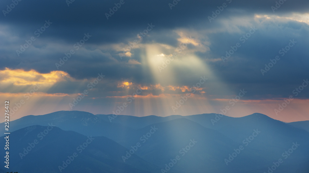 Dramatic sunset in the spring Ukrainian carpathians with red skies, rays and dark clouds, against the background of mountain ridges covered with alpine pine.