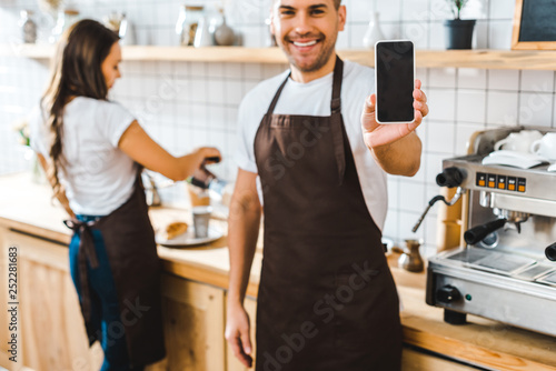 selective focus of handsome cashier showing smartphone with blank screen wile barista working in coffee house