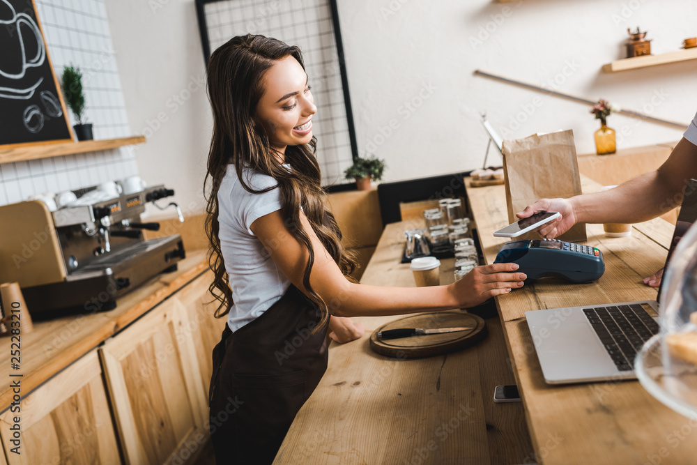 attractive cashier standing in apron and holding terminal wile man paying with smartphone in coffee house attractive cashier standing in apron and holding terminal wile man paying with smartphone in coffee house