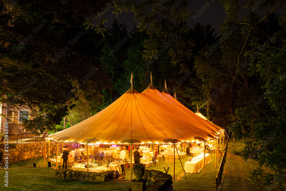 a wedding tent at night surrounded by trees with an orange glow from the lights, long exposure - wedding tent series - obrazy, fototapety, plakaty 