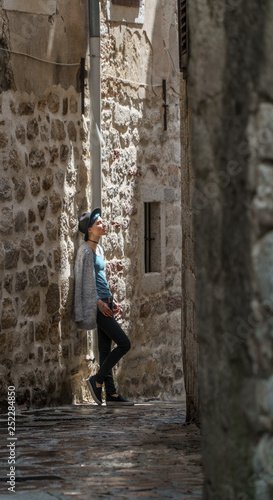 A girl stands in a narrow street in an old European city with a cobbled road and stone walls.