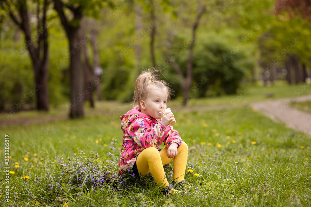 A small cute girl of 3-4 years old is sitting on the grass in early spring and eating a delicious ice cream in a waffle cup. The girl got dirty and dirty.