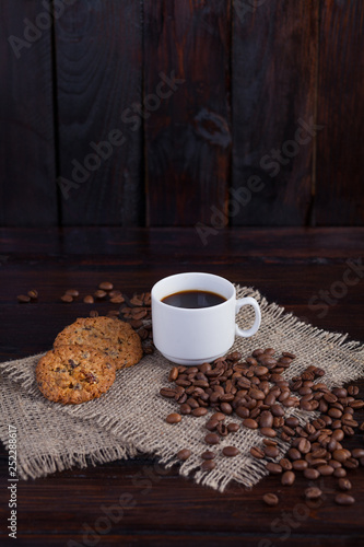 White cup of coffee with coffee grains near and biscuits on linen fabric on the dark vintage background