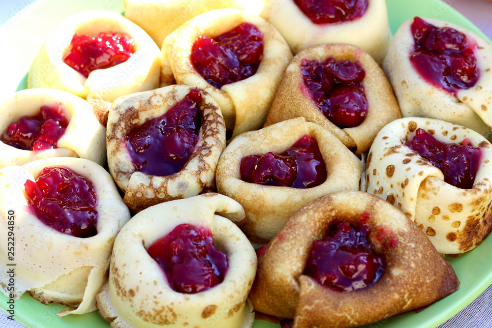 Thin ruddy pancakes on milk with cherry jam, wrapped rolls. Pancakes are packed tightly on a large dish. Pancake week. Pancakes for breakfast and carnival. Homemade baking. Thin ruddy pancakes on milk with cherry jam, wrapped rolls. Pancakes are packed tightly on a large dish. Pancake week. Pancakes for breakfast and carnival. Homemade baking.