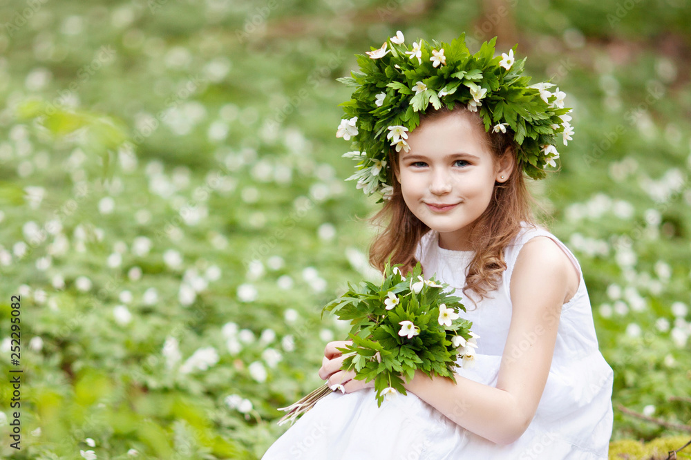 Beautiful little girl in a white dress walks in the spring wood. Portrait of the pretty little girl with a wreath from spring flowers on the head. Easter time. Copy space. Beautiful little girl in a white dress walks in the spring wood. Portrait of the pretty little girl with a wreath from spring flowers on the head. Easter time. Copy space.