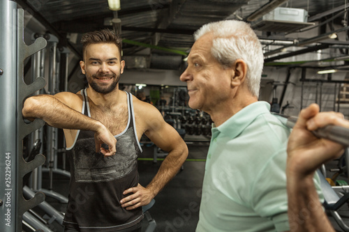 Senior man doing barbell squats with the help of a fitness instructor at the health studio