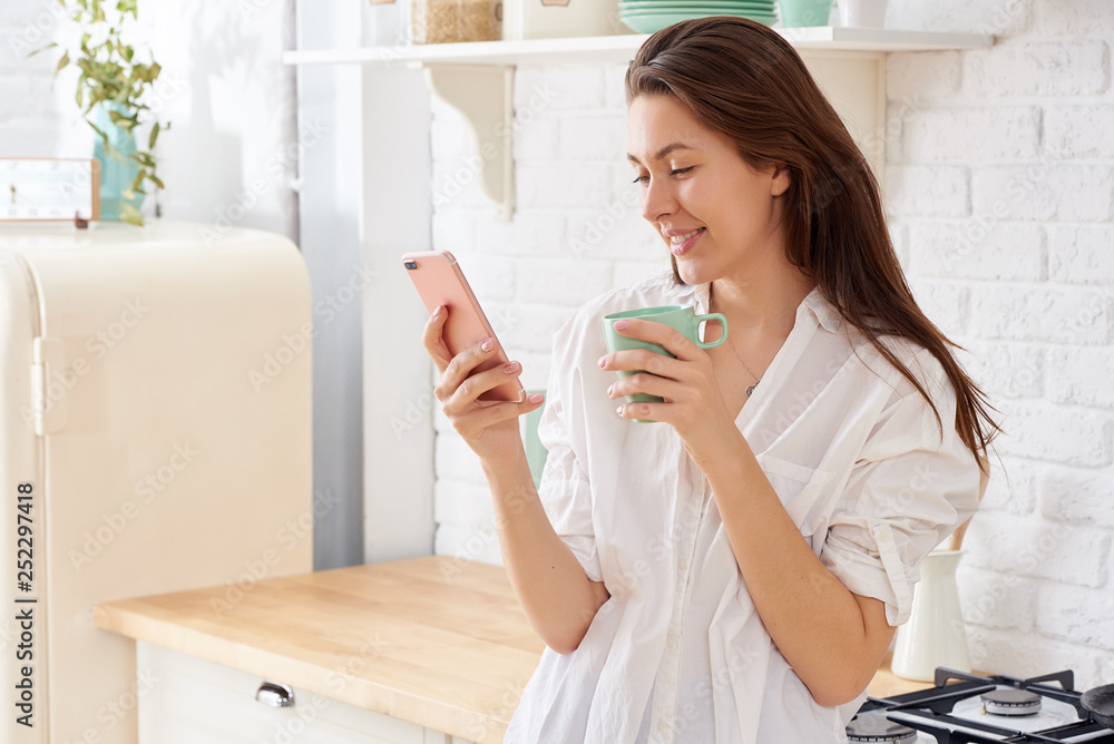Young woman using smartphone leaning at kitchen table with coffee mug and organizer in a modern home. Smiling woman reading phone message. Brunette happy girl typing a text message Young woman using smartphone leaning at kitchen table with coffee mug and organizer in a modern home. Smiling woman reading phone message. Brunette happy girl typing a text message