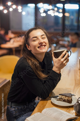 Gorgeous smiling young woman with dark hair eating cake and drinking tea or coffe at a cafeteria in the evening sitting by the window. selective focus  noise effect
