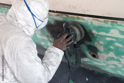 Worker wearing protection suit and mask is sanding down old paint from vessel