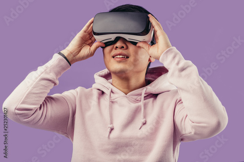 Amazed positive chinese male gamer wearing virtual reality goggles headset, vr box, posing isolated in studio over violet background. Connection, technology, new generation, progress concept.
