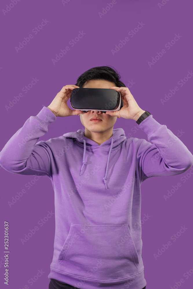 Young Asian guy getting excited during watching 3 D film using virtual reality headset, posing isolated over dark violet background. Positive visualization. Young Asian guy getting excited during watching 3 D film using virtual reality headset, posing isolated over dark violet background. Positive visualization.