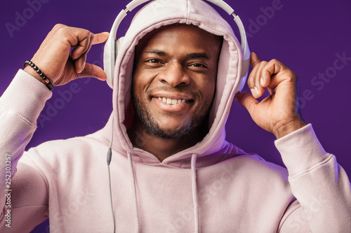 Joyful happy hipster man in white hoodie with hood on listening music with headphones, isolated at purple studio background. Hip hop style, positive emotions, face expression, people concept